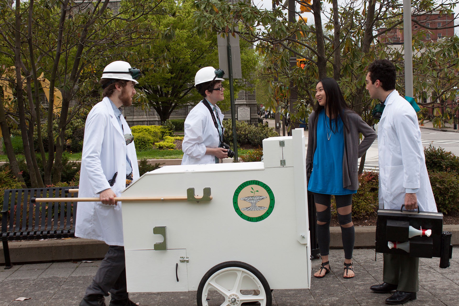 Three people wearing lab coats talking to a women while pushing a white hand cart.