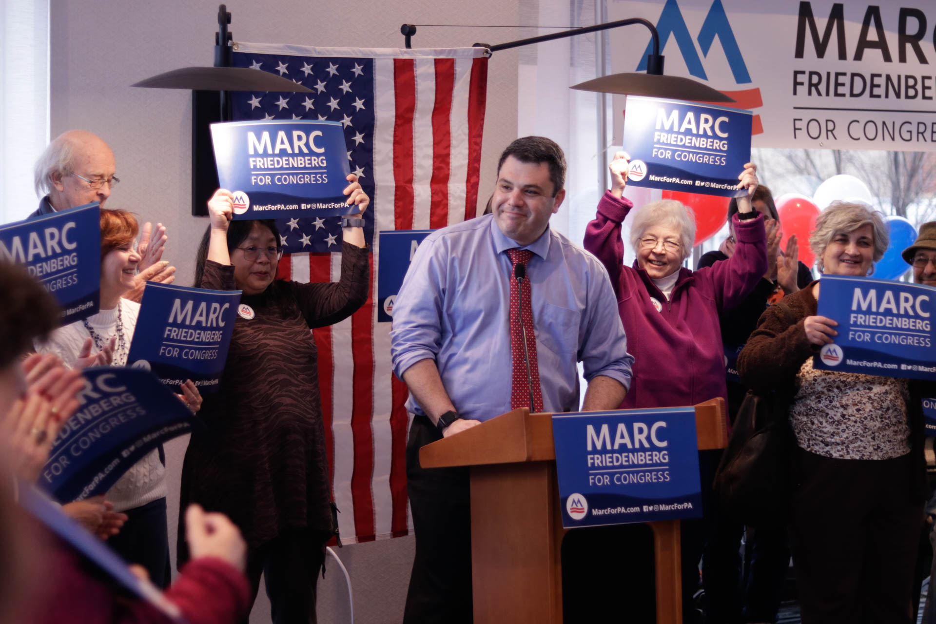 A white man in a tie speaking at a podium, surrounded by people holding signs that say Marc